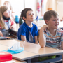 Drei Schulkinder sitzen in in einer Schulklasse an einem Tisch und hören zu.